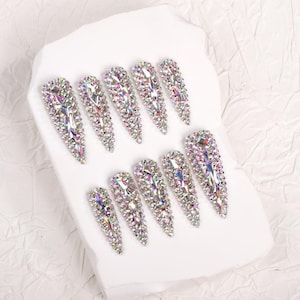 May include: A set of ten long, iridescent, artificial nails. Each nail is covered in small, sparkling, multi-colored rhinestones. The nails are arranged on a white, textured surface.