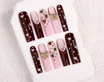 Könnte beinhalten: Ein Set von aufklebbaren Nägeln mit verschiedenen Designs. Einige Nägel sind dunkelbraun mit rosa Herzen, andere sind rosa mit Erdbeeren und goldenen Schleifen, und einige haben herzförmige Edelsteine. Die Nägel sind lang und rechteckig.