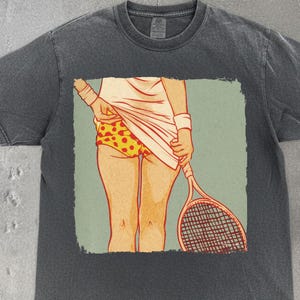 Puede incluir: Camiseta gris oscuro con un gráfico de temática tenística. El gráfico representa a una persona con ropa de tenis sosteniendo una raqueta. La persona lleva una camiseta blanca, pantalones cortos con estampado amarillo y naranja y muñequeras. El diseño está sobre un fondo verde claro.