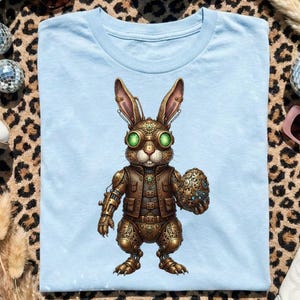 May include: Light blue t-shirt featuring a steampunk rabbit design with goggles and a brown vest, holding a decorative egg. The shirt is surrounded by white cowboy boots, disco balls, sunglasses, and dried grass on a leopard print background.