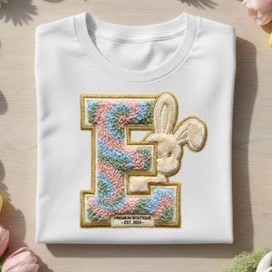 May include: White t-shirt featuring a large letter "E" with a pastel color design and a bunny graphic. The letter is outlined in gold. The text "PREMIUM BOUTIQUE - EST. 2024-" is at the bottom of the letter. Easter theme.