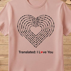 May include: A light pink t-shirt featuring a black heart design made of dashed lines. The text "Translated: I Love You" is printed below the heart. The shirt is surrounded by Valentine's Day decorations.