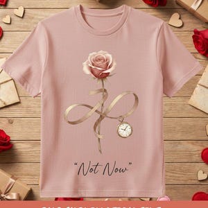 May include: A light pink t-shirt featuring a watercolor rose with a gold ribbon and a pocket watch. The text "Not Now" is printed below the rose. The shirt is surrounded by Valentine's Day decorations.