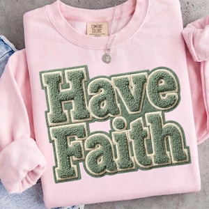 Könnte beinhalten: Ein hellrosa Comfort Colors Sweatshirt mit dem Schriftzug "Have Faith" in einer grünen, strukturierten Schrift im College-Stil. Das Sweatshirt hat einen Rundhalsausschnitt und lange Ärmel. Eine silberne Halskette mit einem kleinen Anhänger ist sichtbar.
