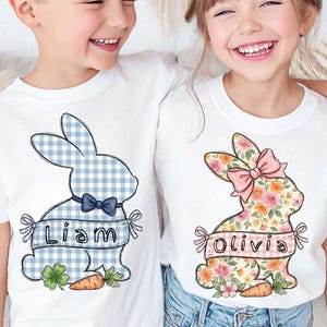 May include: White t-shirts with Easter bunny designs. One bunny is blue gingham with a bow tie and the name "Liam". The other bunny is floral with a pink bow and the name "Olivia". Each design includes a carrot and ribbon.