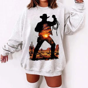 Puede incluir: Una sudadera gris claro con la silueta de un vaquero sosteniendo un lazo contra un fondo de puesta de sol en el desierto. El vaquero lleva sombrero y botas. El diseño incluye cactus y elementos del paisaje desértico.