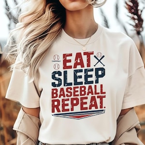 May include: Cream-colored t-shirt with the text "EAT SLEEP BASEBALL REPEAT" in red and navy blue. The design includes baseballs and a baseball bat. The shirt has short sleeves.