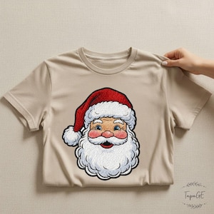 May include: A beige t-shirt featuring a colorful Santa Claus graphic. The Santa illustration has a red hat with white trim, a white beard, rosy cheeks, and blue eyes. The shirt is made of a soft material and is suitable for the holiday season.