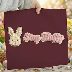 Könnte beinhalten: Ein weinrotes Quadrat mit einem beigen Hasenaufnäher und rosa Text mit der Aufschrift "Stay Fluffy". Der Hasenaufnäher hat ein strukturiertes Aussehen. Der Text ist in einer Schriftart mit hellbeigem Umriss gehalten.