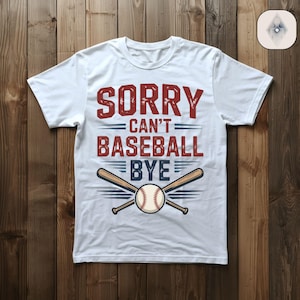 May include: White t-shirt featuring the text "SORRY CAN'T BASEBALL BYE" in red and blue. The design includes crossed baseball bats and a baseball. The shirt is displayed on a wooden surface.