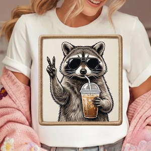 Op de afbeelding: Wit T-shirt met een wasbeer met een zonnebril, een ijskoffie vasthoudt en het vredesteken geeft. De afbeelding is omlijst met een gestikte rand.