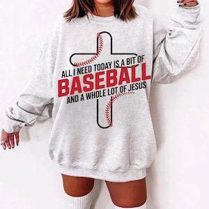 May include: A light gray sweatshirt with the text "ALL I NEED TODAY IS A BIT OF BASEBALL AND A WHOLE LOT OF JESUS" in red and black. The design features a baseball-themed cross.
