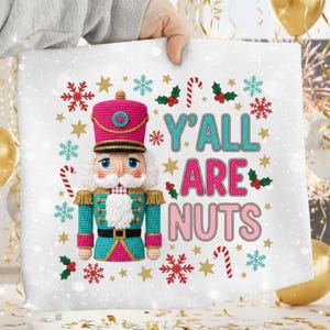 May include: A white square decorative pillow with a crocheted nutcracker design. The nutcracker is wearing a pink hat and teal jacket. The text "Y'ALL ARE NUTS" is displayed with holiday-themed accents.