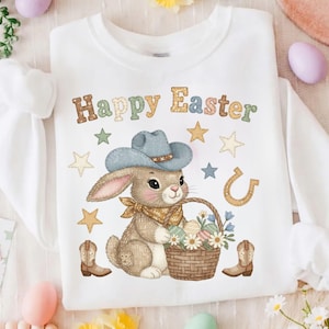 Op de afbeelding: Witte sweatshirt met een "Happy Easter" afbeelding. Het ontwerp toont een konijn met een cowboyhoed en bandana, met een mandje met paaseieren en bloemen. Sterren, een hoefijzer en cowboylaarzen maken het feestelijke ontwerp compleet.
