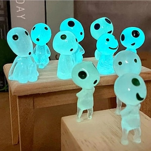 Könnte beinhalten: Eine Sammlung kleiner, durchscheinender, hellblauer, im Dunkeln leuchtender Figuren mit abgerundeten Köpfen und dunklen, kreisförmigen Augen. Die Figuren sind auf Holzoberflächen platziert, wobei einige aufrecht stehen und andere sitzen oder kauern.