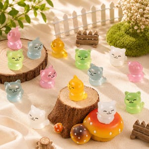 May include: A collection of small, translucent cat figurines in various colours including pink, blue, yellow, green, and white. The cats feature simple, smiling faces and are arranged alongside miniature wooden fence pieces and a mushroom ornament.