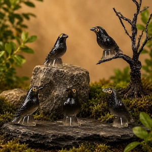 以下が含まれることがあります： 黄色い目をした5羽の小さな黒い鳥の彫刻フィギュアで、それぞれ透明な台座に乗っています。1羽は暗いねじれた枝の上に配置され、残りは大きな質感のある灰色の石の上やその周辺に配置されています。