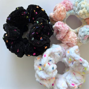 May include: Three colorful scrunchies made of fluffy fabric with confetti-like sprinkles. The scrunchies are black, pink and white.