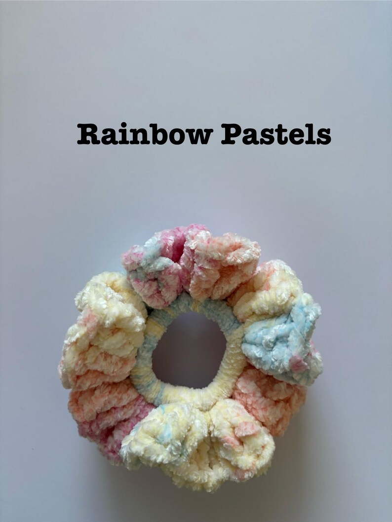 May include: A pastel rainbow colored scrunchie made of fluffy yarn. The scrunchie is in the shape of a donut.