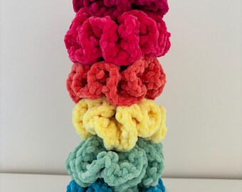 May include: A stack of six colorful fuzzy hair scrunchies in a rainbow gradient. The scrunchies are arranged from top to bottom in the following order: hot pink, pink, orange, yellow, green, and blue.