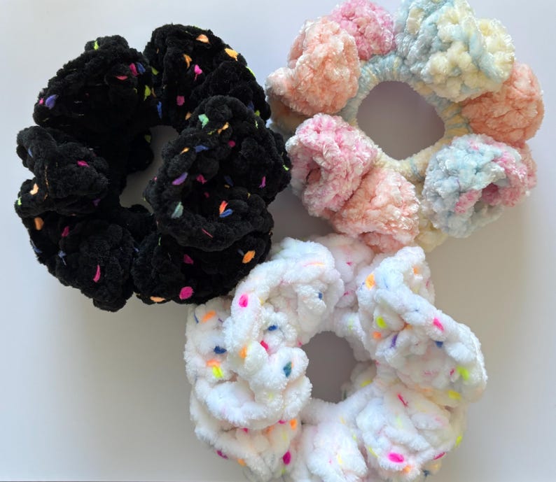 May include: Three colorful scrunchies made of fluffy fabric with confetti-like sprinkles. The scrunchies are black, pink and white.
