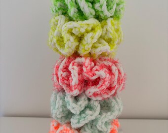 May include: A stack of five colorful crocheted hair scrunchies. The scrunchies are in shades of green, yellow, pink, and orange. They are all made with a fluffy yarn and have a white stripe running through them.