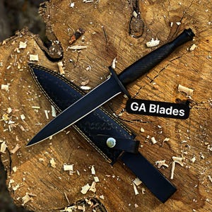 May include: A black dagger with a pointed blade and textured handle, resting on a black leather sheath. The sheath is decorated with a pattern and a strap with a silver button. The text "GA Blades" is visible.