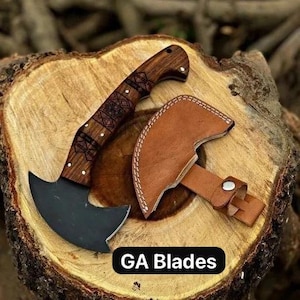 May include: A small, black axe with a wooden handle and decorative carvings, resting on a tree stump. The axe has a leather sheath. The text "GA Blades" is displayed in a black box.