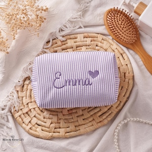 May include: A rectangular cosmetic bag featuring a light purple and white vertical striped pattern. The name Emma is embroidered in purple script on the front, accompanied by a small purple embroidered heart icon to the right of the name.
