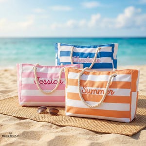 May include: Three striped tote bags in pink, orange, and blue with white stripes, each with a name embroidered on the front. The bags have rope handles and are on a woven mat on a sandy beach. Sunglasses are in front of the pink bag.