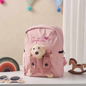 Peut inclure: Sac à dos rose avec le nom "Sophia" brodé en cursive, un cœur rose et un ours en peluche dans une poche en velours côtelé. Le sac a une petite poche latérale et une fermeture éclair noire. Cadeau pour enfants.