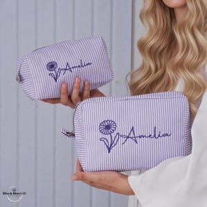 Op de afbeelding: Twee rechthoekige make-uptasjes van lichtpaars en wit gestreepte stof. Elke tas is voorzien van een geborduurde donkerpaarse margriet en de naam Amelia in cursief schrift. Beide tassen hebben een zilverkleurige ritssluiting aan de bovenkant.