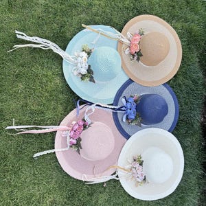 May include: Several wide-brimmed sun hats in shades of light blue, pink, beige, navy, and white. Each hat is decorated with floral arrangements and ribbon ties, set against a backdrop of green grass.