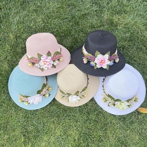 Könnte beinhalten: Sechs Sonnenhüte aus Stroh in verschiedenen Farben, darunter Rosa, Schwarz, Blau, Beige und Weiß. Jeder Hut ist mit einem Blumenschmuck am Rand verziert. Die Hüte liegen auf einer grünen Wiese.