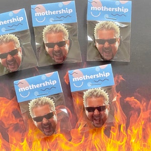 Pode incluir: Cinco sacos de plástico transparentes com etiquetas azuis e cor-de-rosa que dizem "mothership" e contêm uma imagem impressa de um homem com cabelo loiro espetado usando óculos de sol e uma camisa preta. O homem está gritando de boca aberta.
