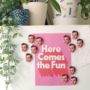 Pode incluir: Um cartaz rosa com o texto "Here Comes the Fun" e v&aacute;rios &iacute;m&atilde;s redondos, coloridos e impressos com o rosto de um homem. Os &iacute;m&atilde;s est&atilde;o presos a uma superf&iacute;cie branca.