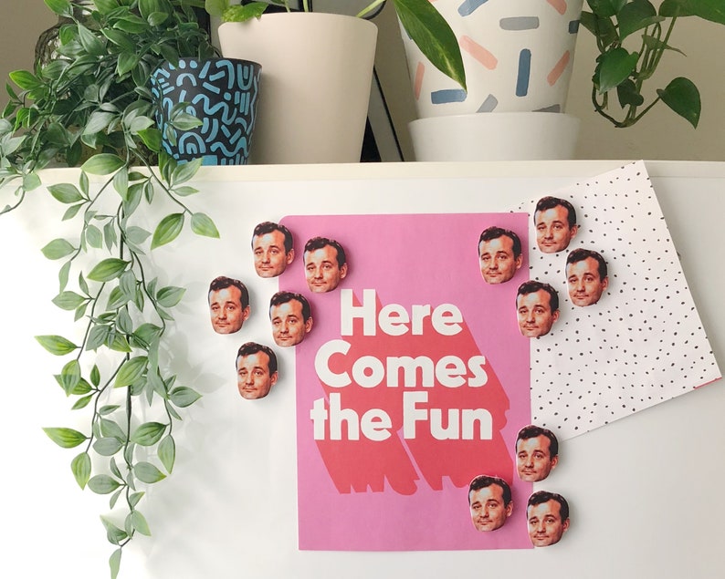 Pode incluir: Um cartaz rosa com o texto "Here Comes the Fun" e v&aacute;rios &iacute;m&atilde;s redondos, coloridos e impressos com o rosto de um homem. Os &iacute;m&atilde;s est&atilde;o presos a uma superf&iacute;cie branca.