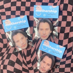 Pode incluir: Tr&ecirc;s sacos de pl&aacute;stico transparentes com etiquetas azuis e cor-de-rosa que dizem "mothership" e apresentam um rosto sorridente. Cada saco cont&eacute;m um bot&atilde;o redondo com uma imagem de Keanu Reeves.