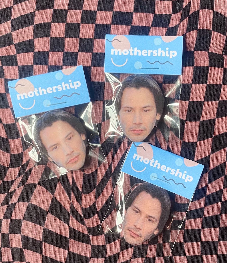 Pode incluir: Tr&ecirc;s sacos de pl&aacute;stico transparentes com etiquetas azuis e cor-de-rosa que dizem "mothership" e apresentam um rosto sorridente. Cada saco cont&eacute;m um bot&atilde;o redondo com uma imagem de Keanu Reeves.