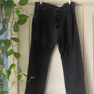 May include: A pair of black denim jeans hanging on a wooden hanger. The jeans have a straight leg and a button fly.