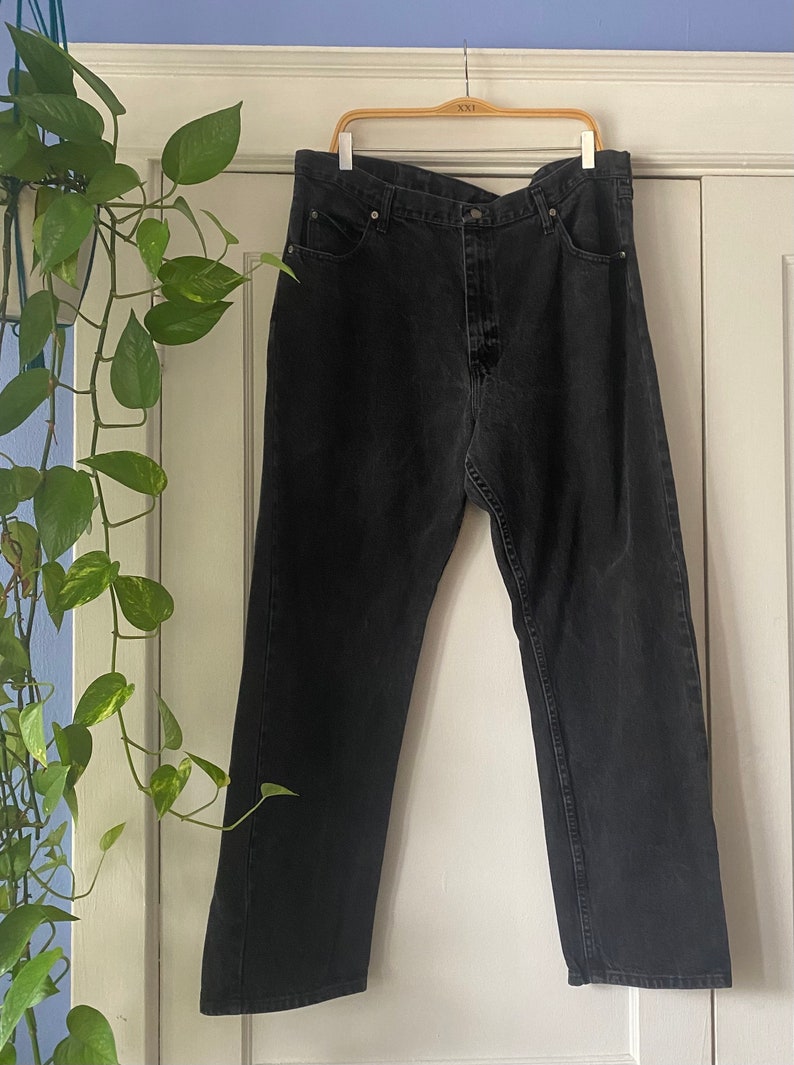 May include: A pair of black denim jeans hanging on a wooden hanger. The jeans have a straight leg and a button fly.