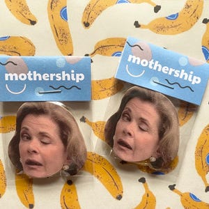 Könnte beinhalten: Zwei Ansteckbuttons mit dem Gesicht einer Frau mit einem geschlossenen Auge. Jeder Button ist mit einer hellblauen Karte verpackt, auf der "mothership" in weißer Schrift steht. Der Hintergrund ist ein Muster aus gelben Bananen.