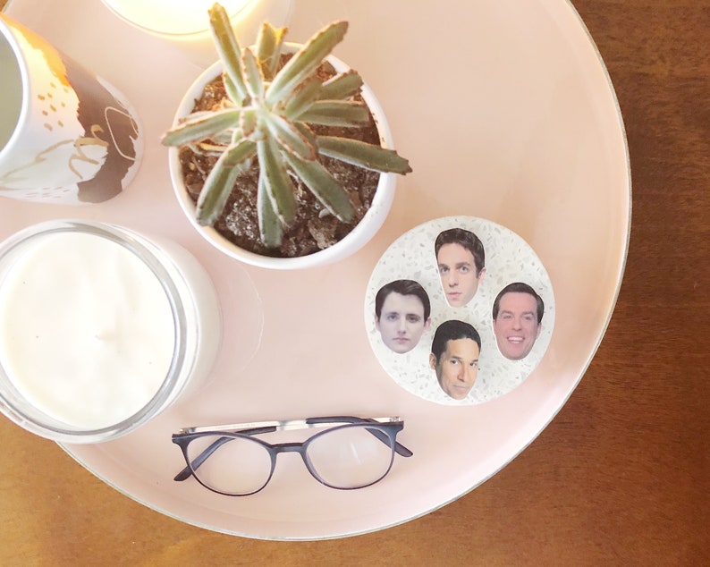 Pode incluir: Uma bandeja rosa com uma vela branca, uma planta suculenta em um vaso branco, &oacute;culos e um descanso de copo branco com quatro rostos impressos nele.