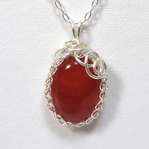 May include: A silver chain necklace with a red carnelian gemstone pendant. The pendant is set in a silver wire cage with a delicate swirl design.