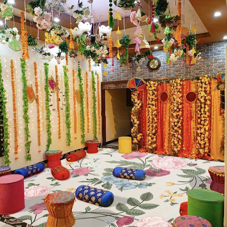 May include: A floral decorated room with a white floral patterned carpet. The room is decorated with yellow and orange flower garlands, green garlands, and white string lights. The room has a backdrop of orange and red fabric with yellow and orange flower garlands. There are several small, round, colorful stools and pillows on the floor.