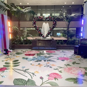 May include: A white floor with a floral pattern in pink, blue, green, and yellow. A heart-shaped floral arrangement is in the background with a fountain in the center. The floor is decorated for a wedding or other special event.