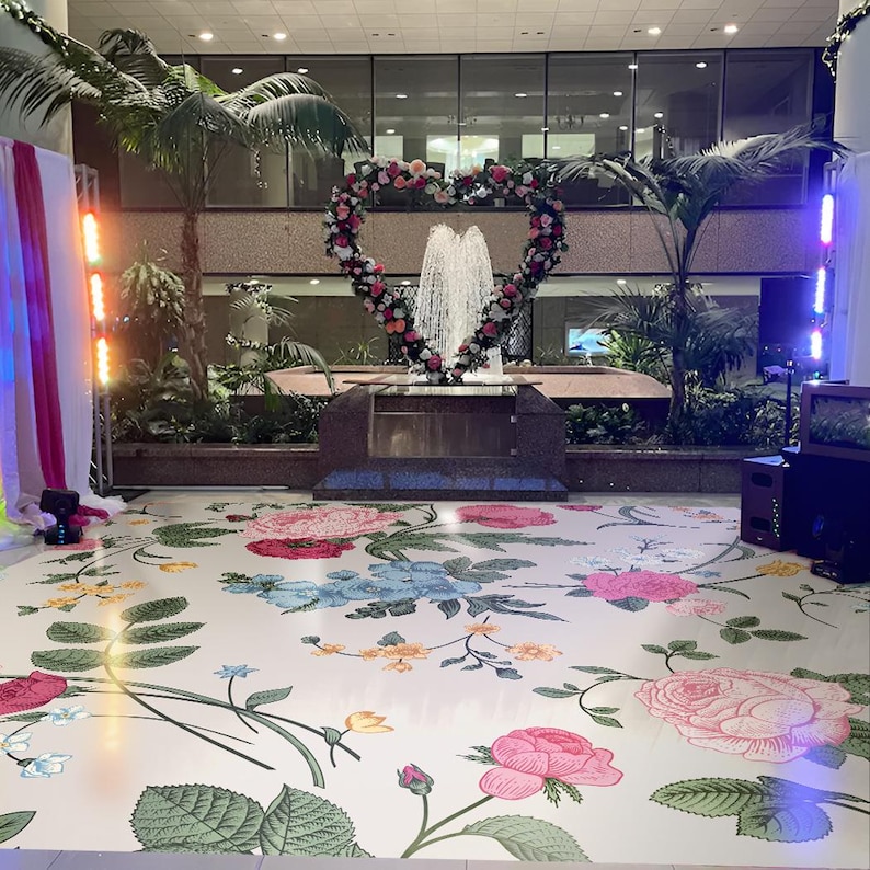 May include: A white floor with a floral pattern in pink, blue, green, and yellow. A heart-shaped floral arrangement is in the background with a fountain in the center. The floor is decorated for a wedding or other special event.