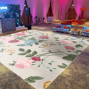 May include: A white floral patterned rug with pink, yellow, blue, and green flowers. The rug is laid out on a light brown floor in a large room with white curtains and pink lighting.