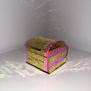 Peut inclure: Un petit coffre décoratif avec un couvercle bombé. Le coffre est recouvert de petits carreaux carrés dorés, avec des bandes de carreaux roses. Le coffre est posé sur une surface blanche.