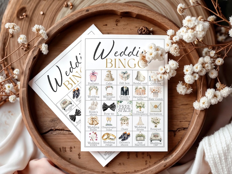 May include: Two wedding bingo cards with illustrations of wedding-related items like a cake, rings, and a dress. The cards are on a wooden tray with white flowers and the text "Wedding BINGO".